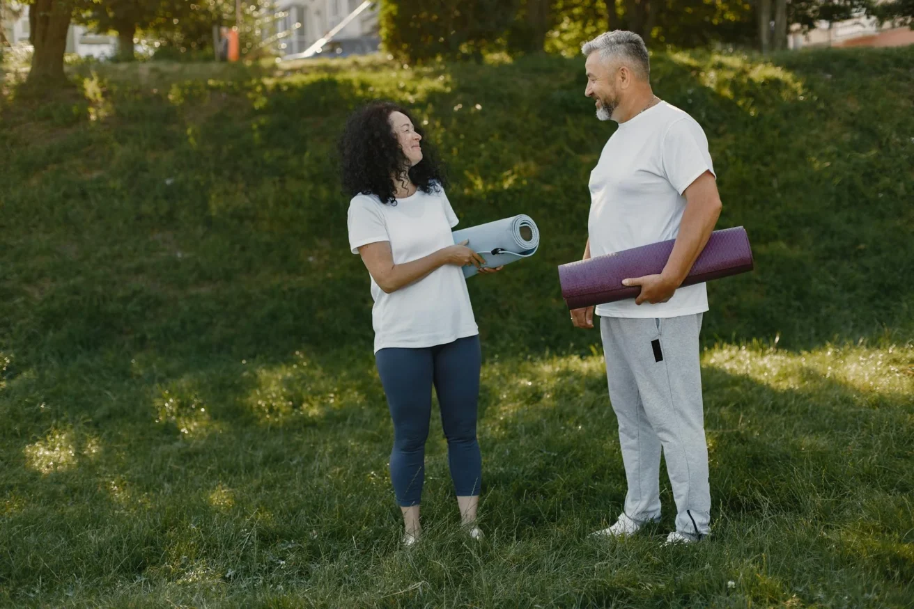 Two adults, a woman and a man, stand on grass outdoors holding yoga mats and smiling at each other, both dressed in casual athletic wear with trees in the background.