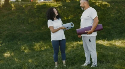 Two adults, a woman and a man, stand on grass outdoors holding yoga mats and smiling at each other, both dressed in casual athletic wear with trees in the background.