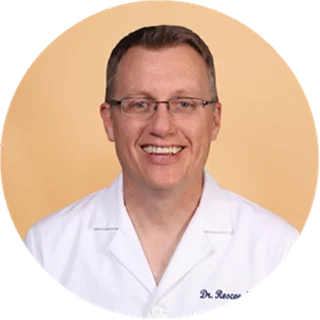A smiling man wearing glasses and a white lab coat stands against a plain light peach background.
