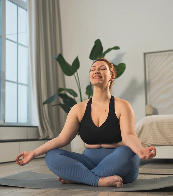 A woman in a black sports bra and blue leggings sits cross-legged on a yoga mat indoors, smiling and meditating with her hands resting on her knees. There is a bed and a large plant in the background.