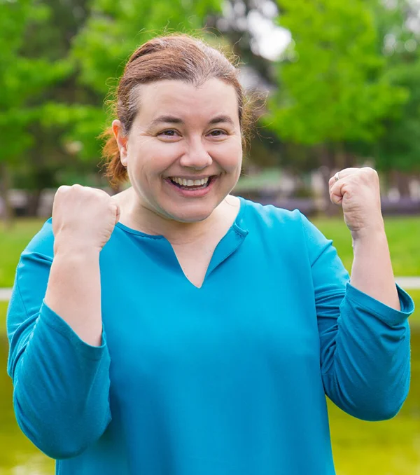 A woman in a teal top smiles widely outdoors, holding both fists up in a celebratory gesture. Green trees and blurred nature are visible in the background.