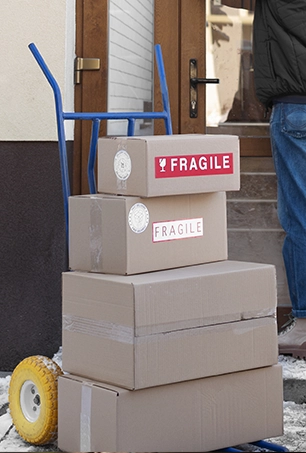 A dolly with four stacked cardboard boxes, two labeled fragile, stands near a building entrance. A person in jeans and a jacket is partially visible by the door.