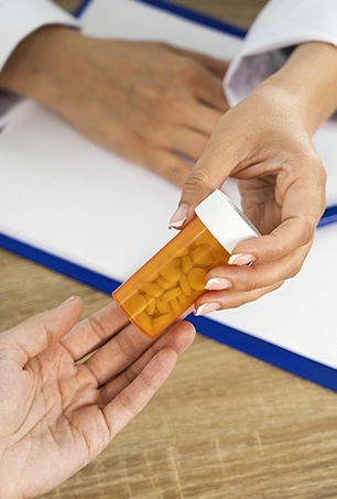 A person hands an orange prescription pill bottle to another person over a wooden desk, with a clipboard and paper in the background.