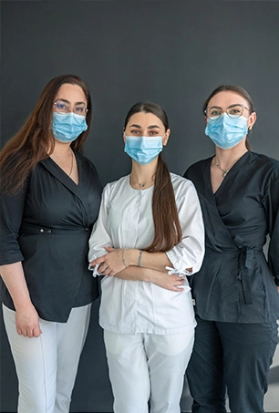 Three women wearing medical masks and glasses stand together against a plain dark background. Two wear black tops and light pants, while the woman in the center wears a white medical uniform.