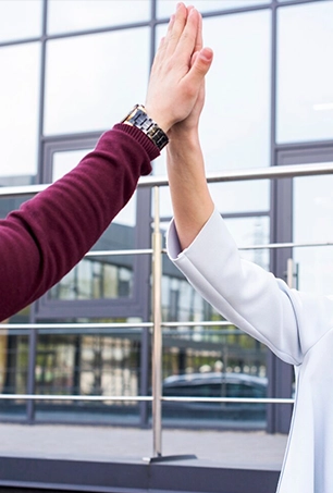 Two people giving a high five outdoors in front of a modern glass building. Only their arms are visible; one person wears a maroon sleeve and a watch, the other wears a white sleeve.