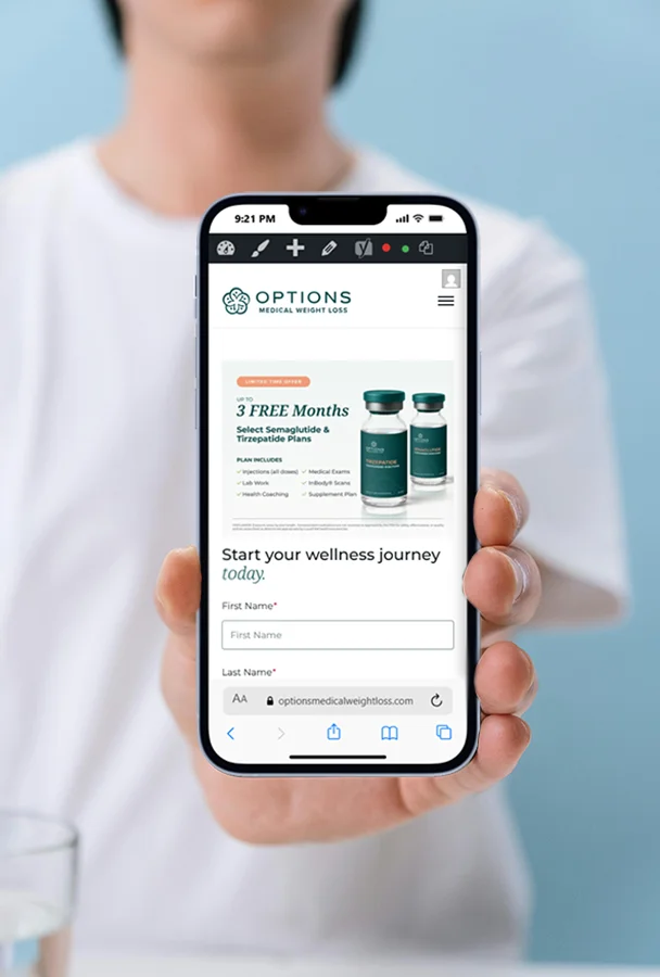 A person holds a smartphone displaying the Options Medical Weight Loss website, which advertises three free months of select semaglutide and tirzepatide treatments with an image of medication bottles and a wellness journey sign-up form.
