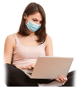A woman wearing a face mask sits cross-legged and types on a laptop, appearing focused on her work.