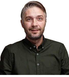 A man with short, light brown hair and a trimmed beard is smiling slightly. He is wearing a dark green button-up shirt and is posed against a brown, blurred background.