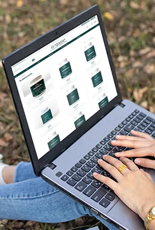 A person using a laptop outdoors sits on grass, browsing a website displaying various product containers. The screen shows a grid layout of items. The person types on the keyboard with both hands visible.