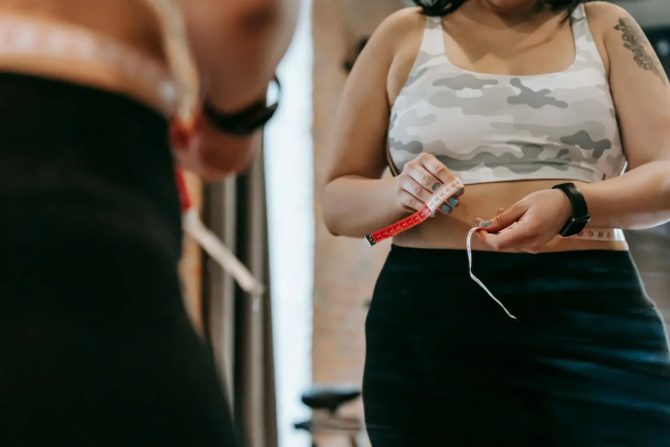 A woman in workout clothes stands in front of a mirror, holding a measuring tape around her waist, focusing on tracking her fitness progress. Her reflection is partially visible.
