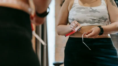 A woman in workout clothes stands in front of a mirror, holding a measuring tape around her waist, focusing on tracking her fitness progress. Her reflection is partially visible.