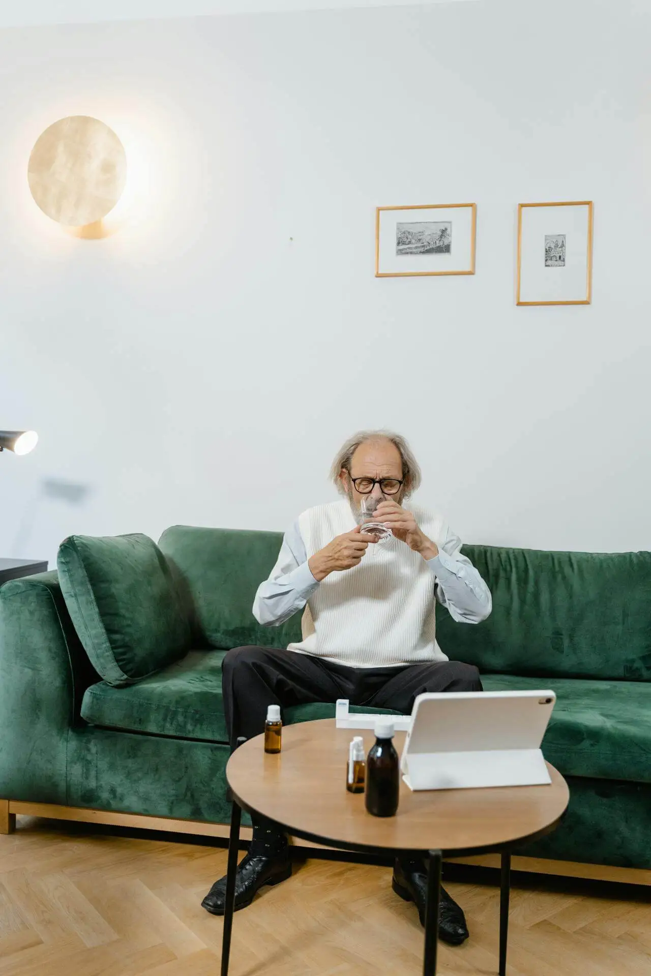 An older man sits on a green sofa, wearing glasses and holding a glass of water, with medicine bottles and a tablet on a round wooden table in front of him, in a bright living room.