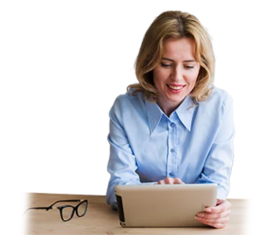 A woman with blonde hair, wearing a light blue shirt, sits at a wooden table using a tablet. A pair of black eyeglasses rests on the table beside her. She is smiling and appears focused on the device.