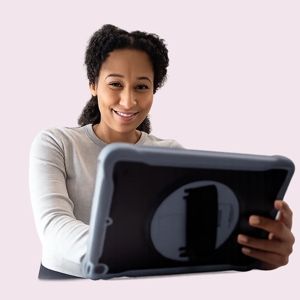 A woman with curly hair smiles while holding and looking at a tablet device against a light pink background.