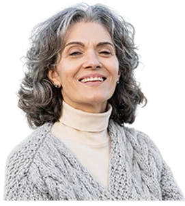 A smiling older woman with gray, wavy hair wearing a light-colored turtleneck and a knitted sweater, standing outside with a blurred background.