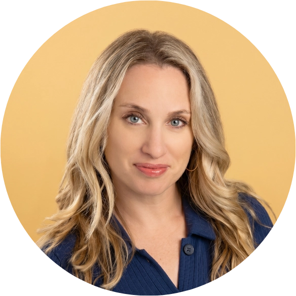 A woman with long, wavy blonde hair and blue eyes, wearing a navy blue top, poses in front of a solid light yellow background. She is facing the camera and smiling softly.