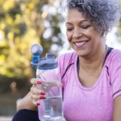 A smiling woman with short curly gray hair sits outdoors, holding a clear water bottle. She wears a pink athletic shirt and has red painted nails. Trees and greenery are blurred in the background.